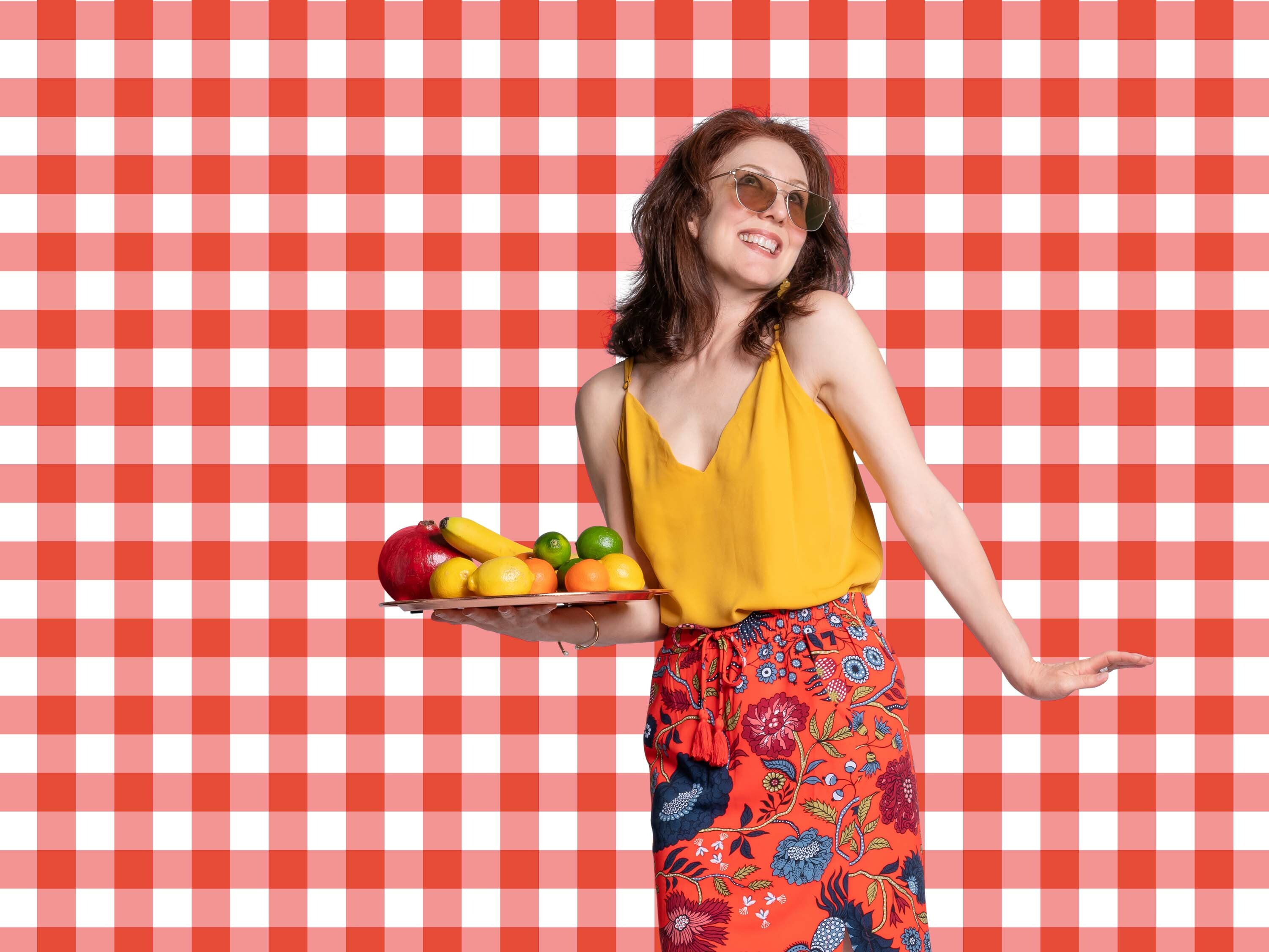 Woman in yellow top floral skirt holding platter of fruit against red checkered background communal living recipes