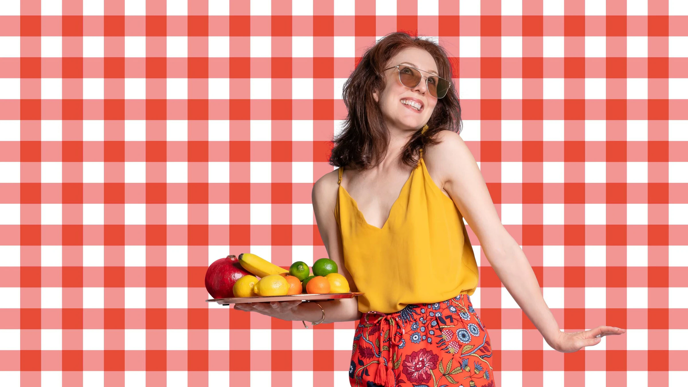 Woman in yellow top floral skirt holding platter of fruit against red checkered background communal living recipes